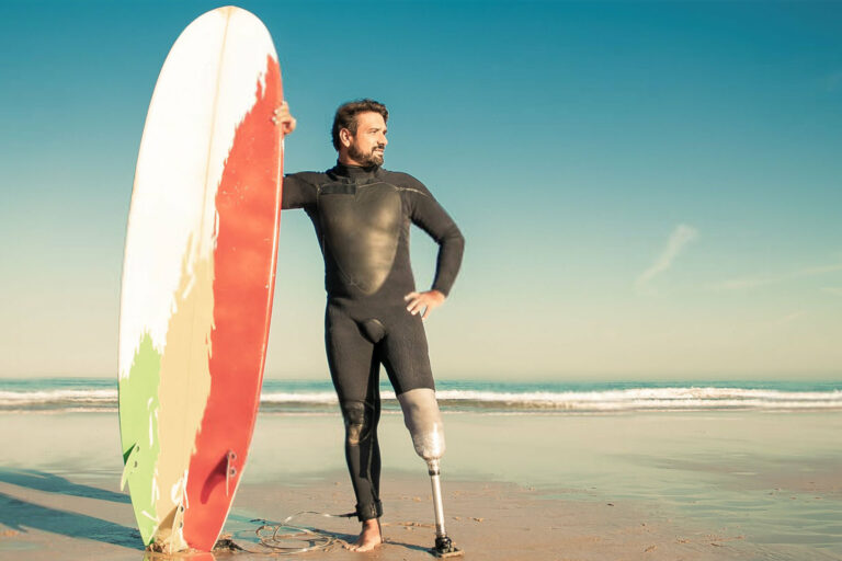 Confident handicapped man standing on sea beach with board. Attractive brunette man with artificial leg wearing black wetsuit and looking at ocean. Physical disability and extreme sport concept